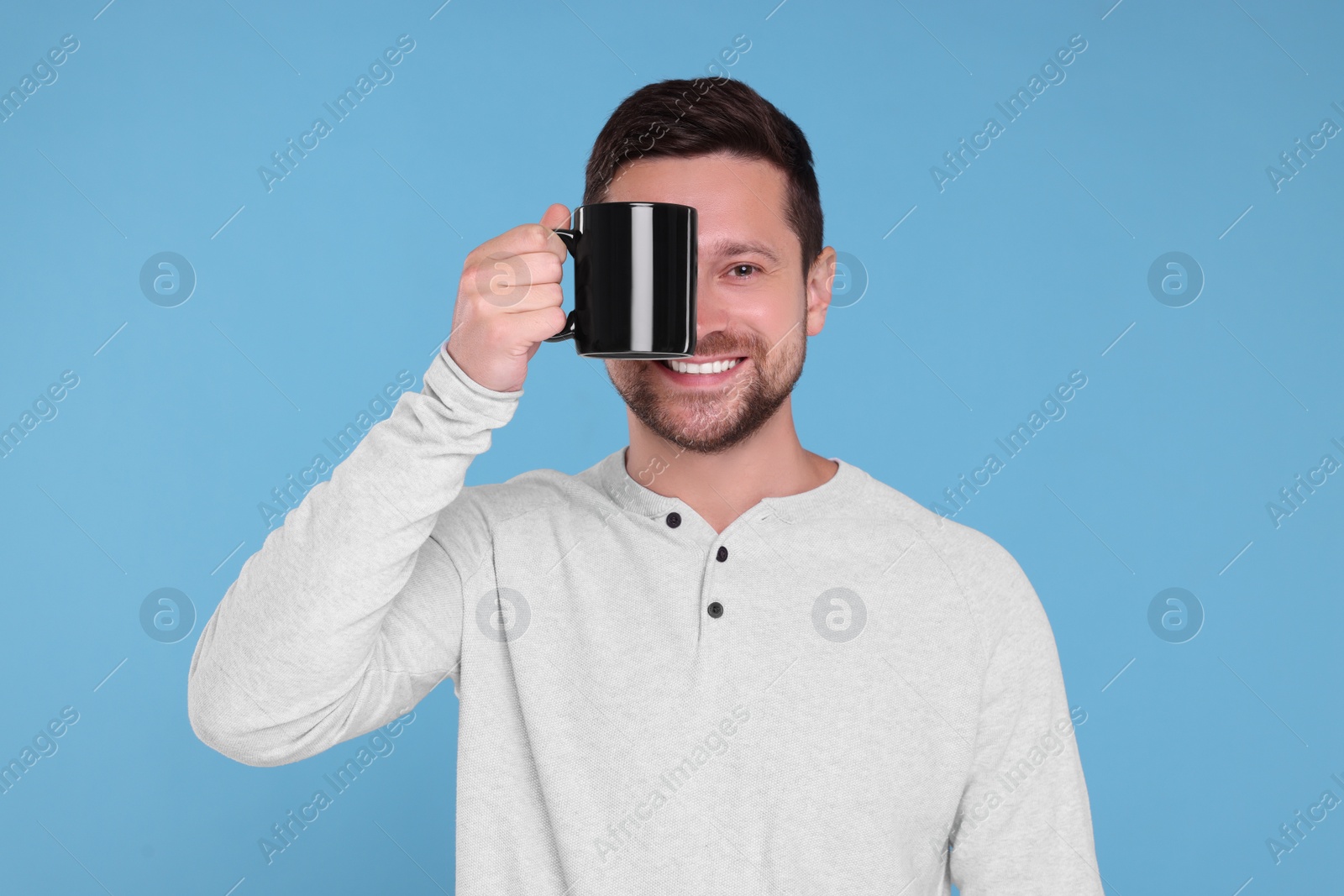 Photo of Portrait of happy man covering eye with black mug on light blue background