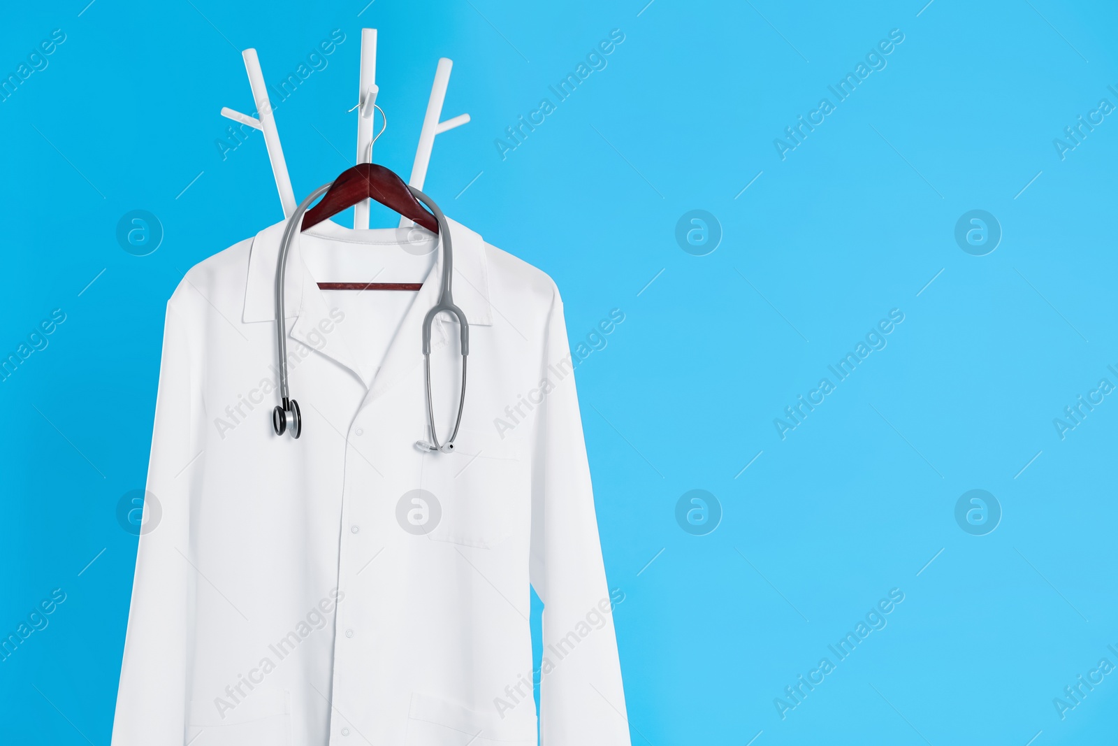 Photo of White medical uniform and stethoscope hanging on rack against light blue background. Space for text