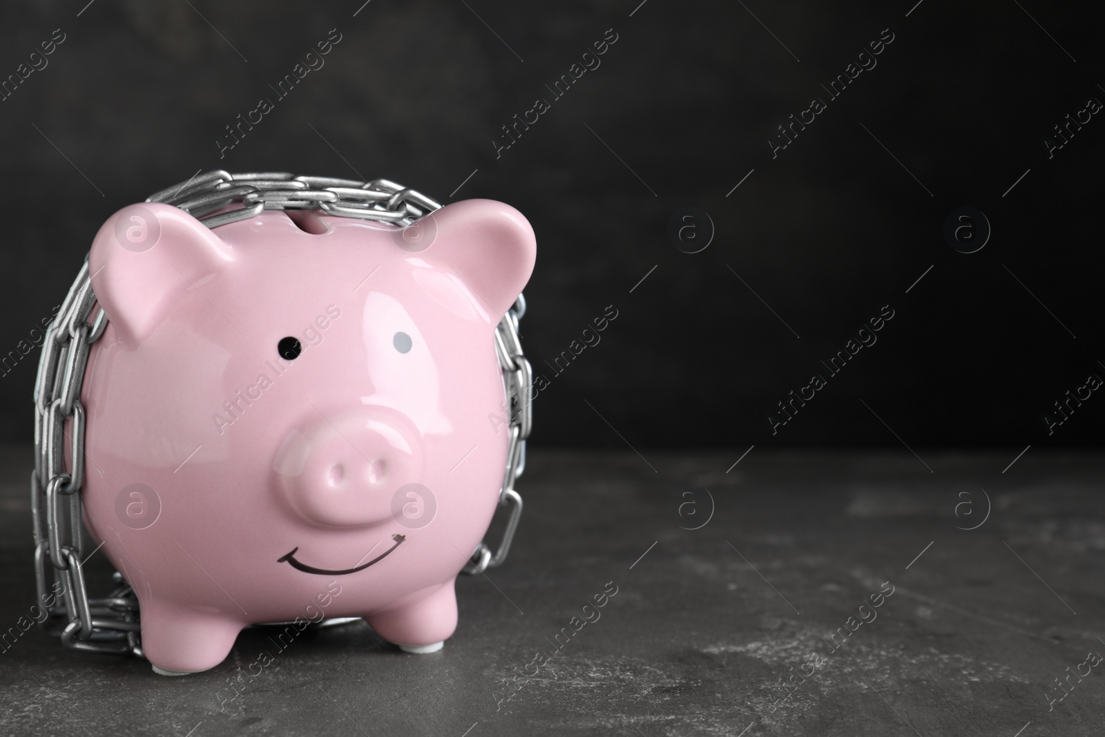 Photo of Piggy bank with steel chain on dark grey stone table, space for text. Money safety concept