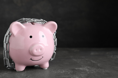 Photo of Piggy bank with steel chain on dark grey stone table, space for text. Money safety concept
