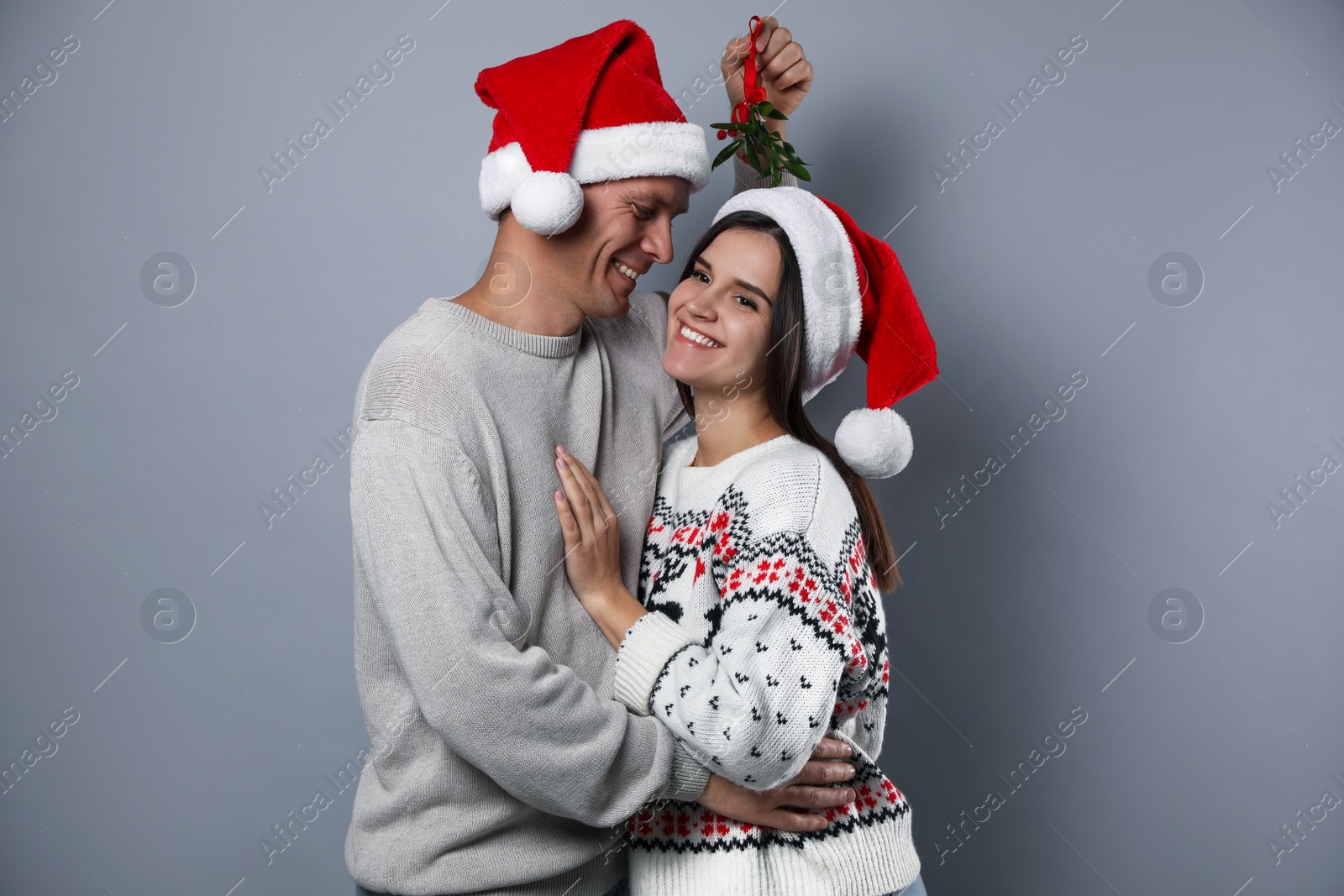 Photo of Happy couple in Santa hats standing under mistletoe bunch on grey background