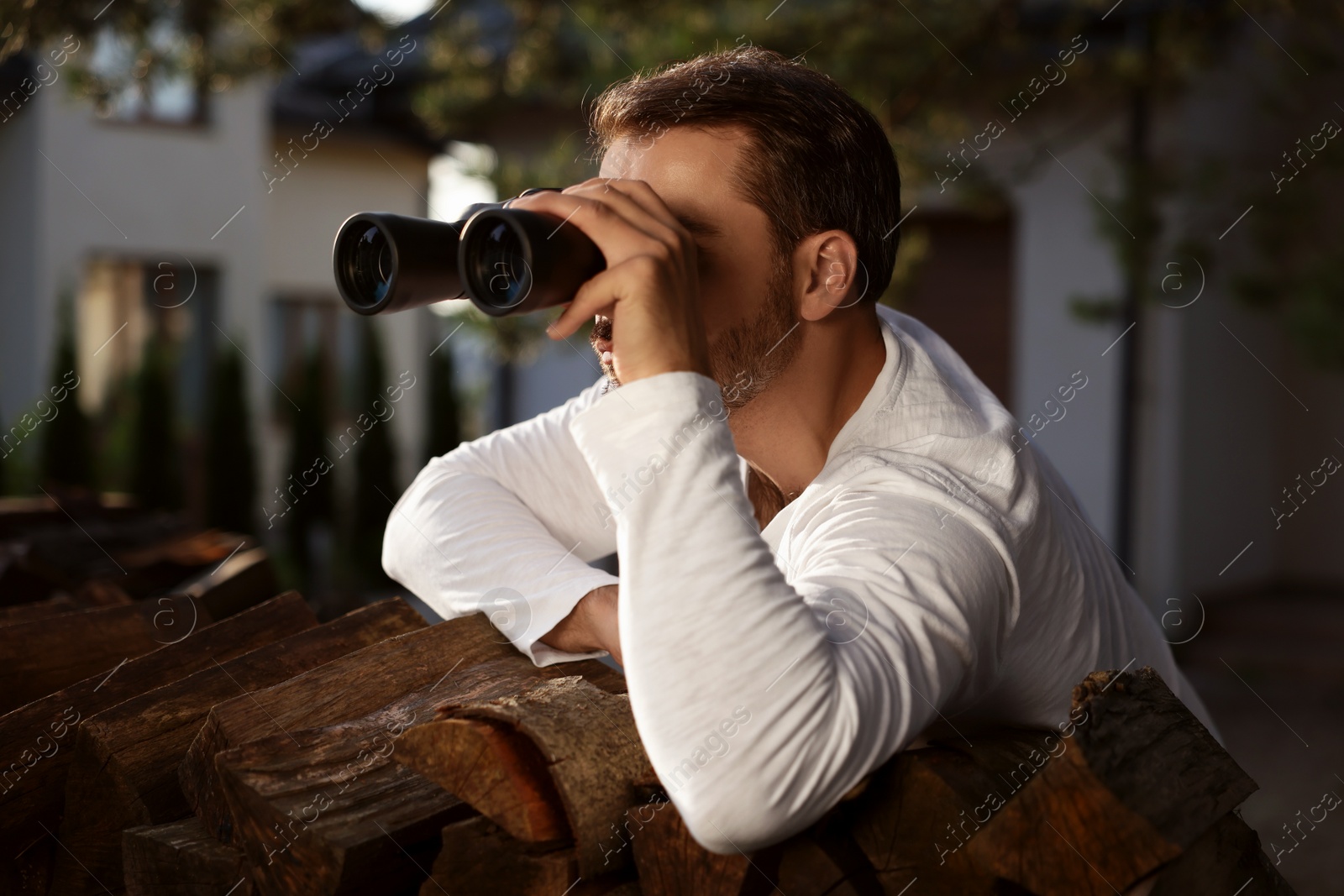 Photo of Concept of private life. Curious man with binoculars spying on neighbours over firewood outdoors