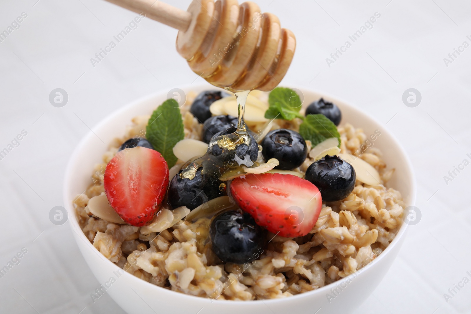 Photo of Pouring honey onto tasty oatmeal with strawberries, blueberries and almond petals in bowl on white tiled table, closeup