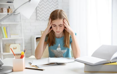 Photo of Teenage girl suffering from headache while doing homework at table indoors