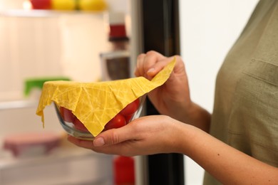 Woman taking away beeswax food wrap from bowl of fresh tomatoes near refrigerator in kitchen, closeup