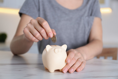 Photo of Woman putting money into piggy bank at marble table indoors, closeup