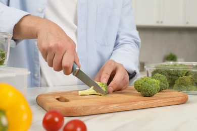 Photo of Man cutting fresh broccoli with knife near glass container at white marble table in kitchen, closeup. Food storage