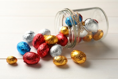 Photo of Overturned glass jar with chocolate eggs wrapped in colorful foil on white wooden table