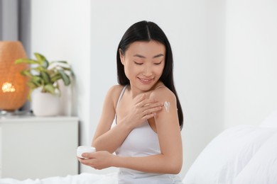 Photo of Beautiful young Asian woman applying body cream on shoulder in bedroom