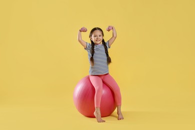 Cute little girl with dumbbells and fit ball on yellow background