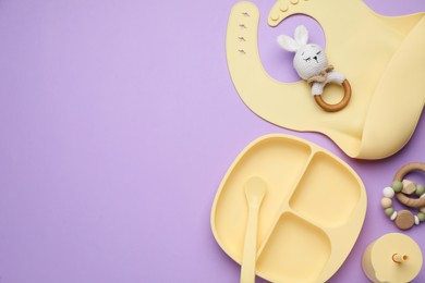 Photo of Set of plastic dishware, silicone bib and baby accessories on violet background, flat lay. Space for text