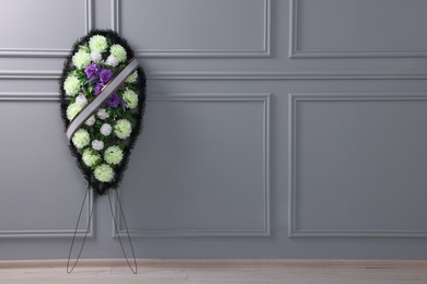 Photo of Funeral wreath of plastic flowers with ribbon near light grey wall indoors, space for text