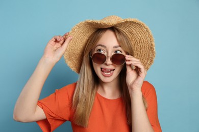 Beautiful young woman wearing straw hat and sunglasses on light blue background. Stylish headdress