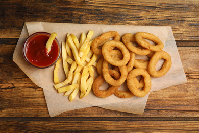 Delicious onion rings, fries and ketchup on wooden table, top view