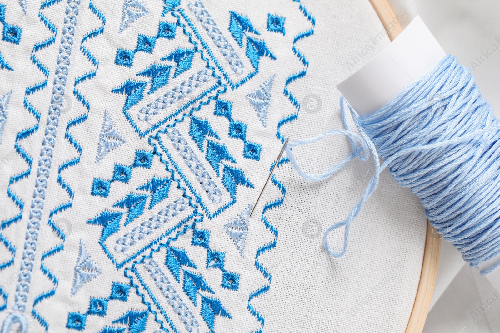 Photo of White fabric with blue Ukrainian national embroidery in hoop, needle and thread, top view