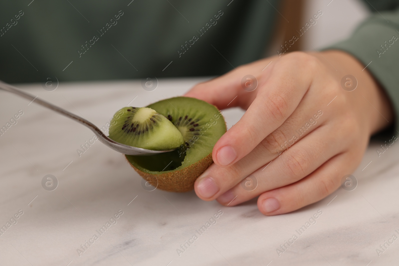 Photo of Boy eating tasty fresh kiwi with spoon at white marble table indoors, closeup