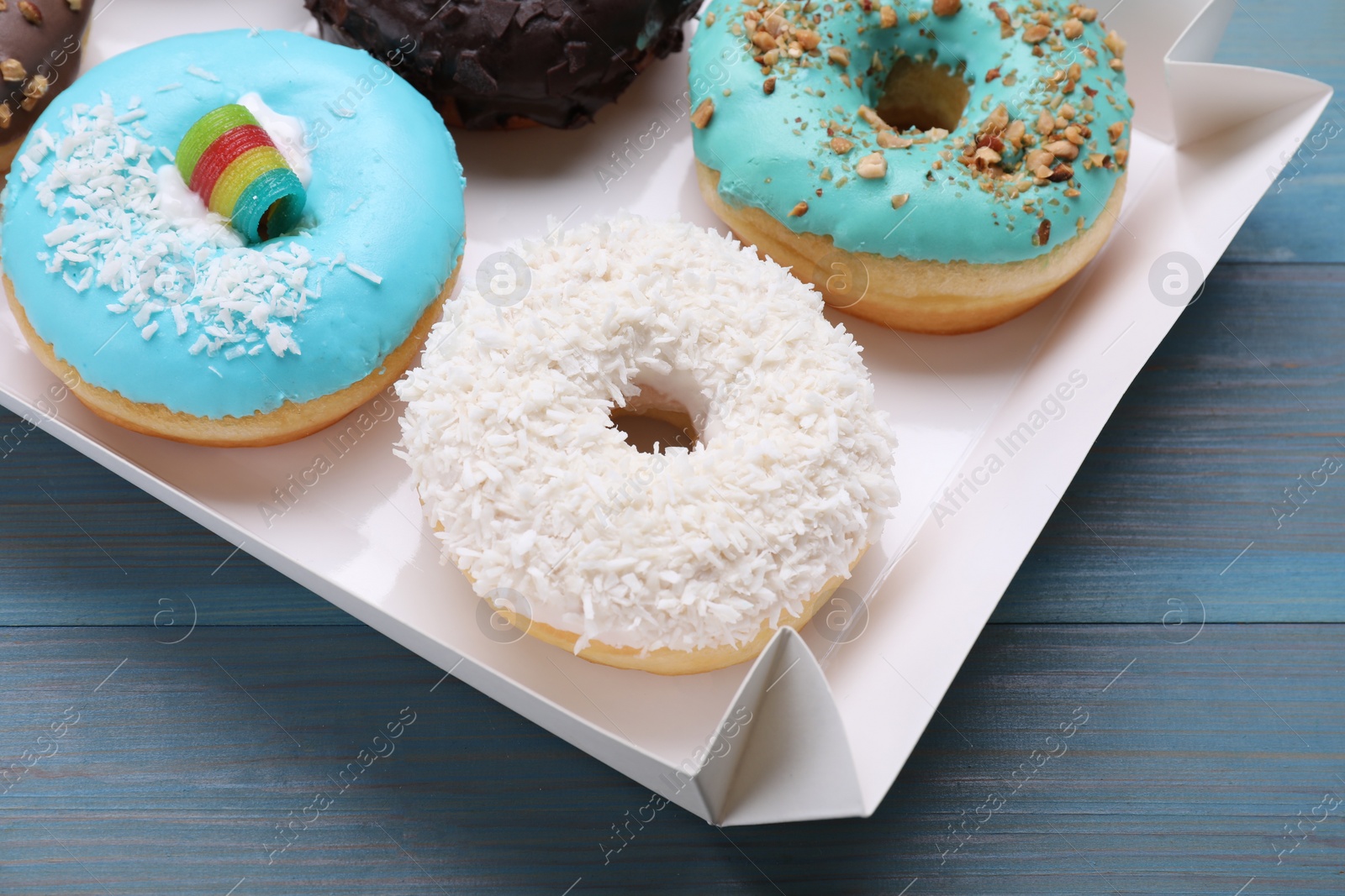 Photo of Box with different tasty glazed donuts on light blue wooden table, closeup