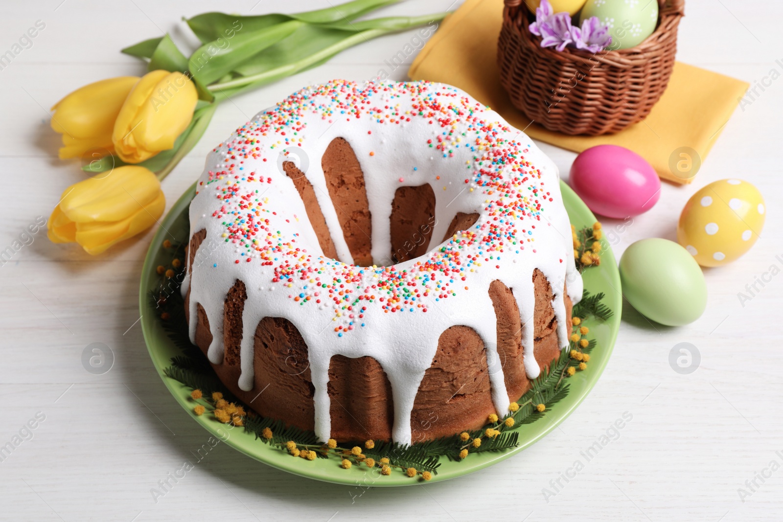 Photo of Glazed Easter cake with sprinkles, painted eggs and flowers on white wooden table