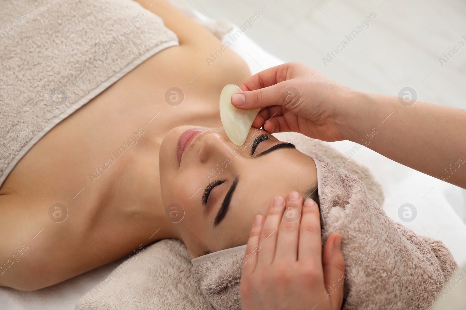 Photo of Young woman receiving facial massage with gua sha tool in beauty salon