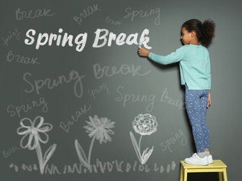 Image of African American girl writing phrase Spring Break with chalk on grey wall. School holiday