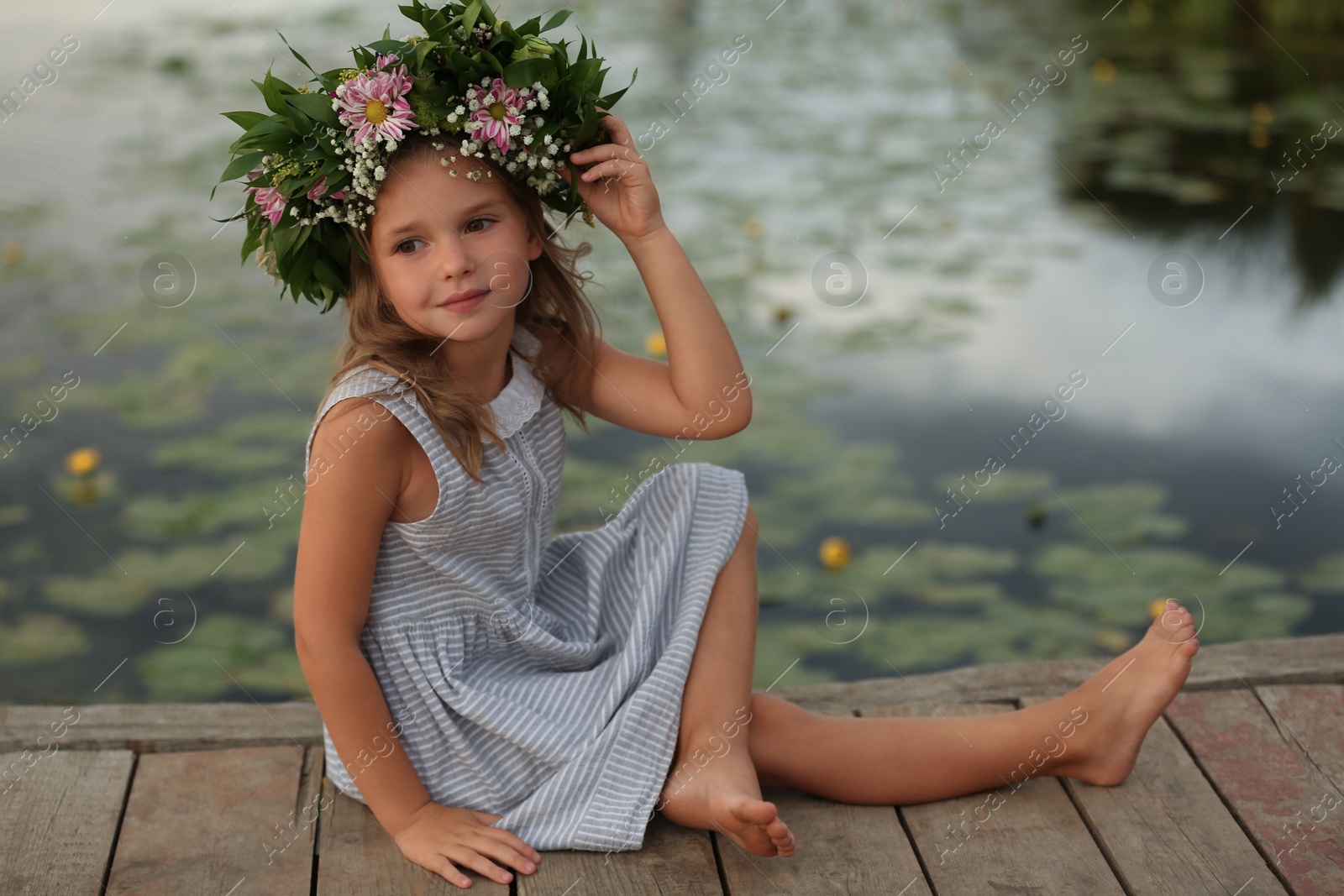 Photo of Cute little girl wearing wreath made of beautiful flowers on pier near pond