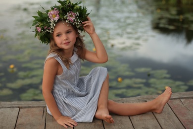 Photo of Cute little girl wearing wreath made of beautiful flowers on pier near pond