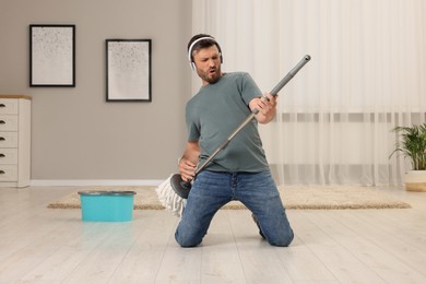 Photo of Enjoying cleaning. Happy man in headphones with mop singing while tidying up at home