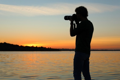 Young male photographer taking photo of riverside sunset with professional camera outdoors