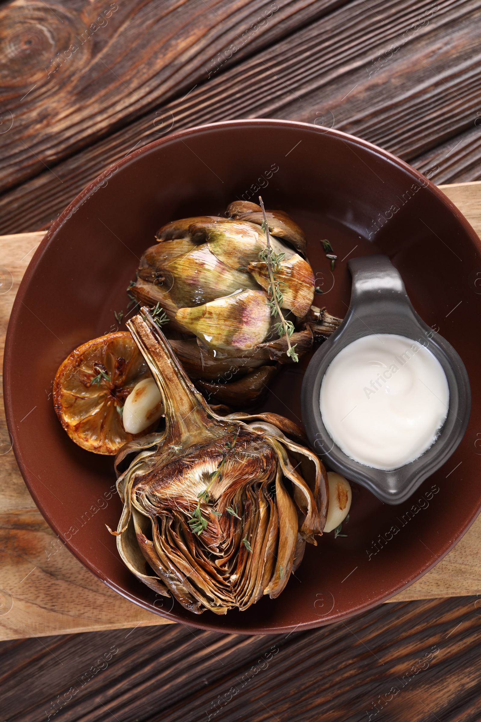 Photo of Tasty grilled artichoke and sauce in bowl on wooden table, top view