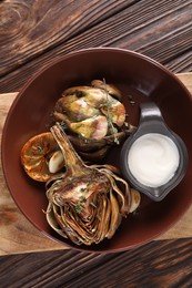 Tasty grilled artichoke and sauce in bowl on wooden table, top view