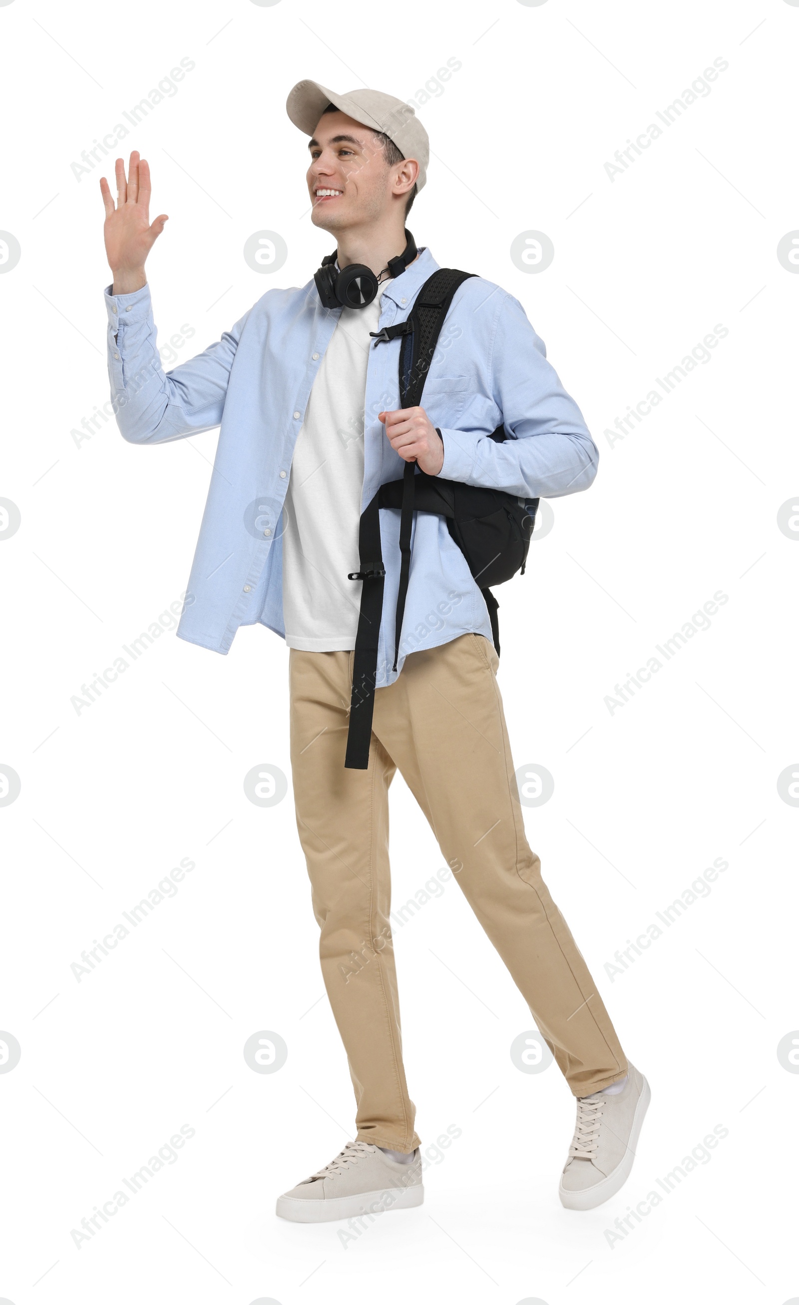 Photo of Young man with cap, headphones and backpack waving hand on white background