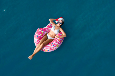 Image of Young woman wearing Santa hat and bikini with inflatable ring in sea, top view. Christmas vacation