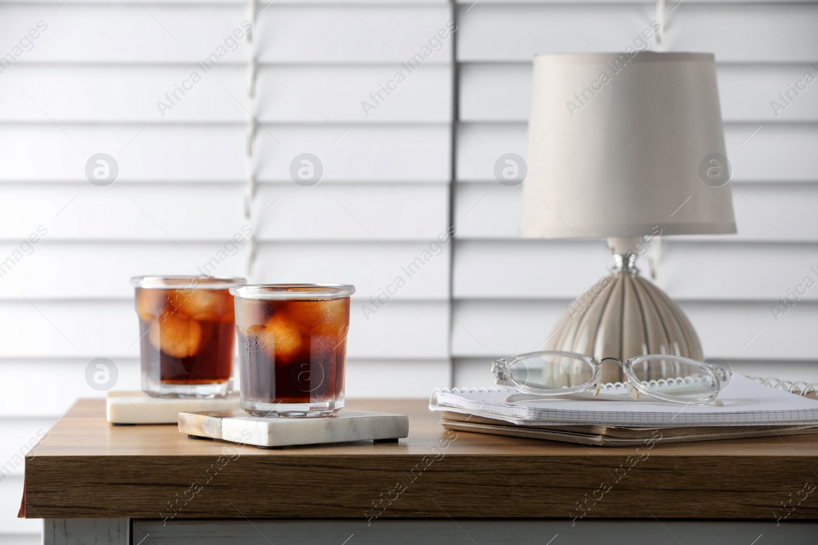 Photo of Glasses of cold drink with stylish cup coasters, lamp and stationery on wooden table in room