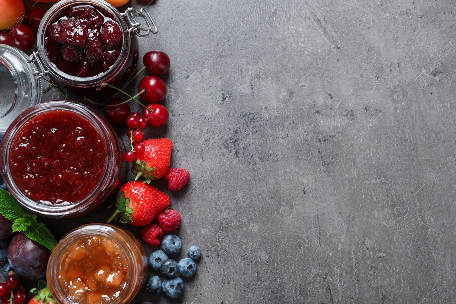 Photo of Jars with different jams and fresh fruits on grey table, flat lay. Space for text