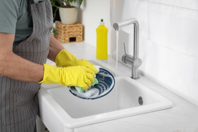 Man in protective gloves washing plate above sink in kitchen, closeup