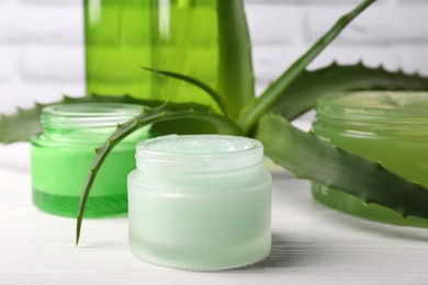 Photo of Jars of cosmetic gel and aloe vera leaves on white wooden table, closeup