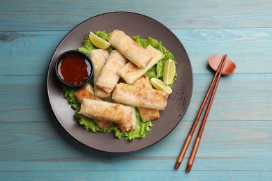 Photo of Plate with tasty fried spring rolls, lettuce, lime, sauce and chopsticks on light blue wooden table, top view