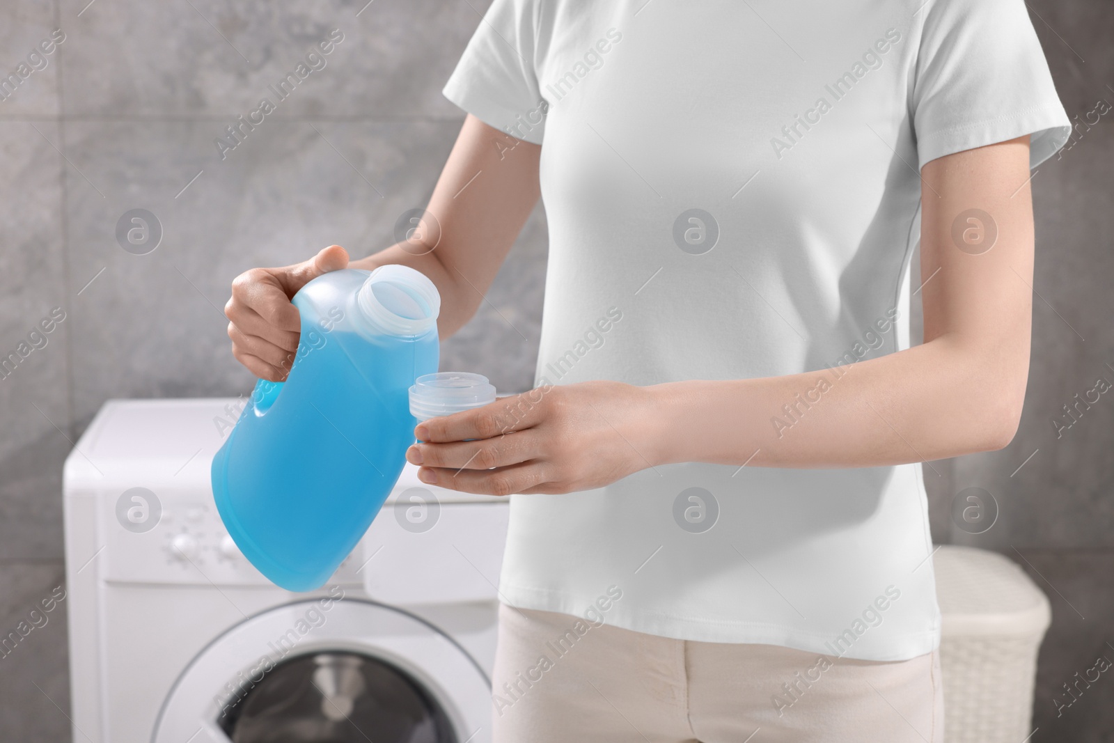 Photo of Woman pouring fabric softener from bottle into cap for washing clothes indoors, closeup