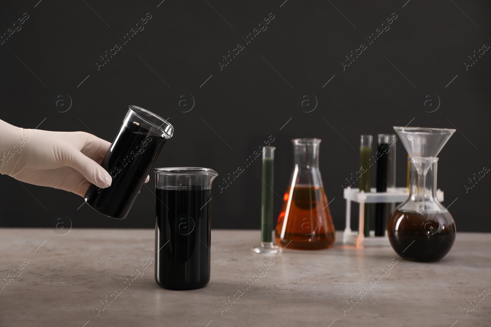 Photo of Woman pouring black crude oil into beaker at grey table against dark background, closeup