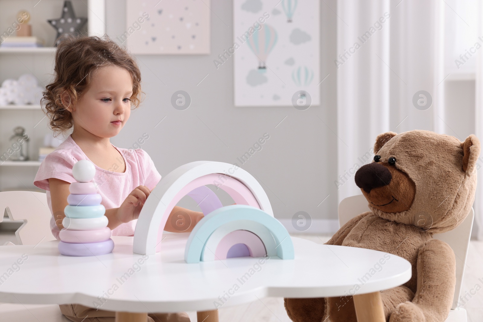 Photo of Cute little girl playing with toy and teddy bear at white table in room