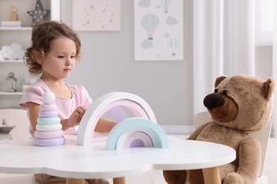 Cute little girl playing with toy and teddy bear at white table in room