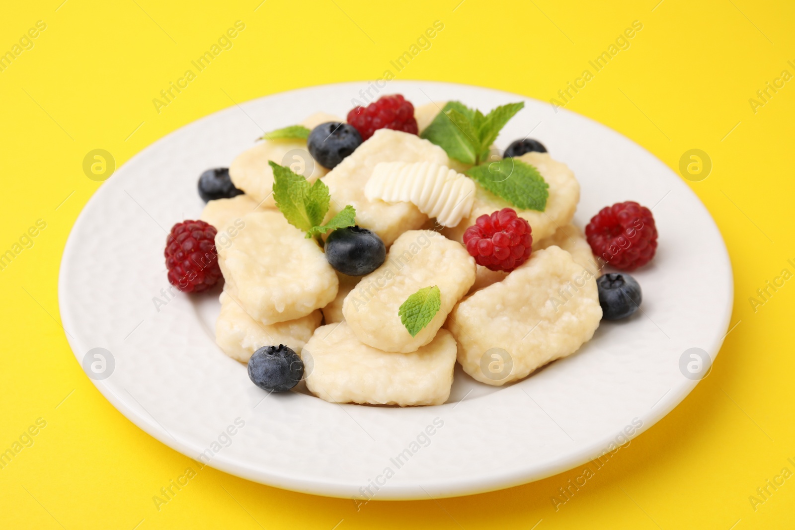 Photo of Plate of tasty lazy dumplings with berries, butter and mint leaves on yellow background