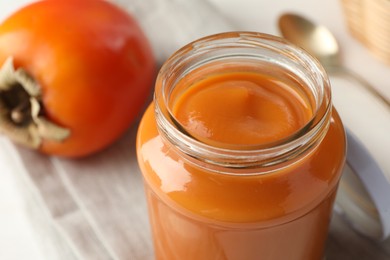 Photo of Delicious persimmon jam in glass jar and fresh fruit on table, closeup