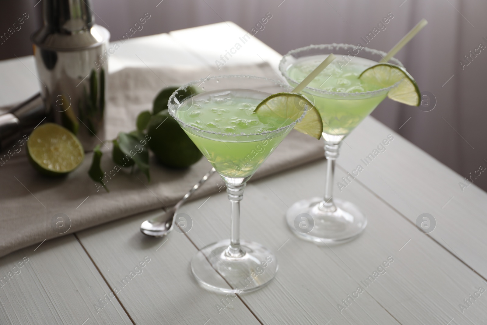 Photo of Delicious Margarita cocktail in glasses, limes and bartender equipment on white wooden table