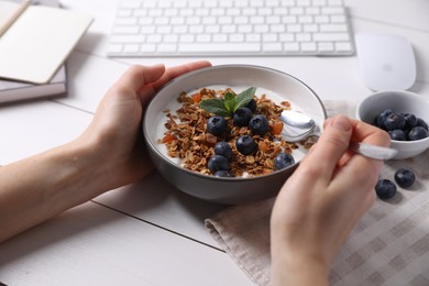 Woman holding bowl of tasty granola with blueberries at white wooden table, closeup