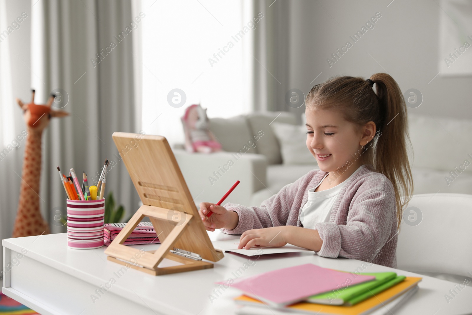 Photo of Adorable little girl doing homework with tablet at table indoors