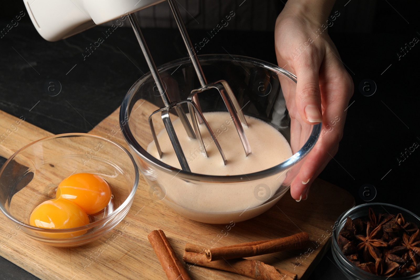 Photo of Woman whipping ingredients with mixer at black table, closeup. Cooking delicious eggnog