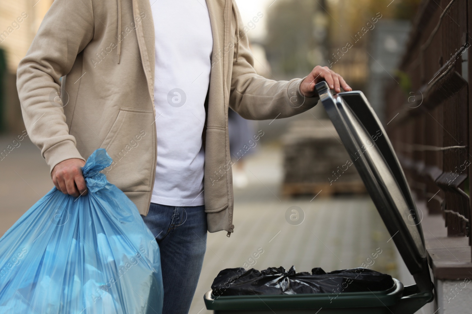 Photo of Man putting garbage bag into recycling bin outdoors, closeup