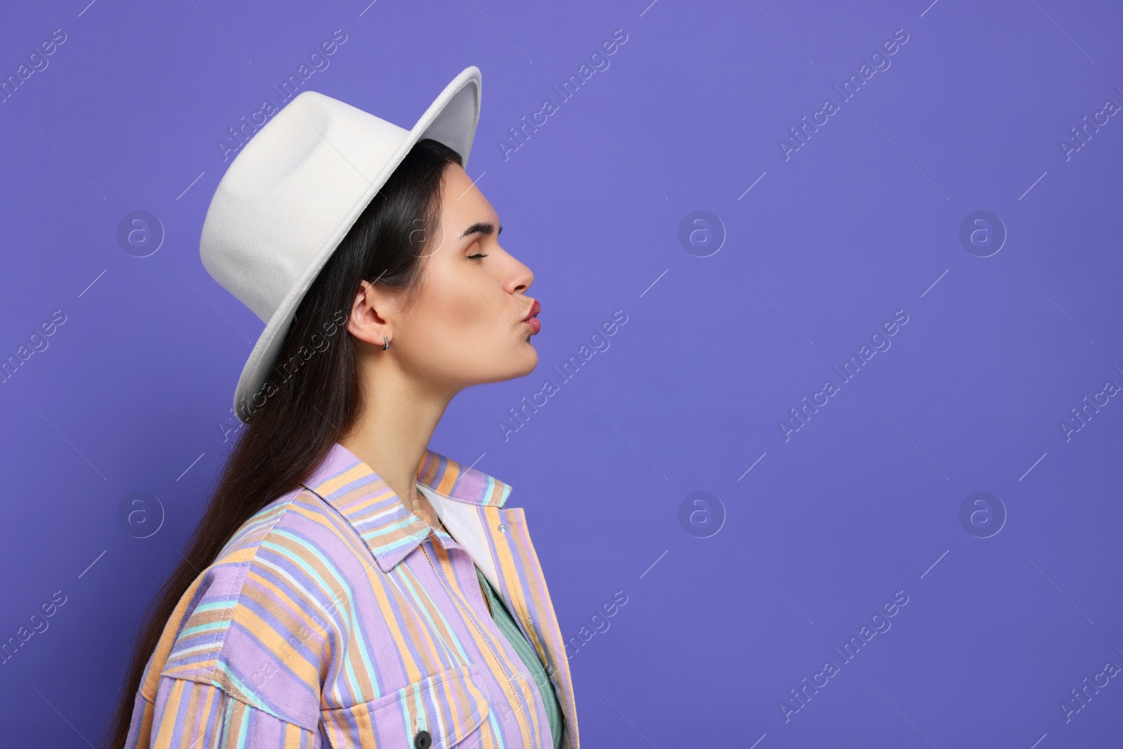 Photo of Beautiful young woman with stylish hat giving kiss on purple background. Space for text