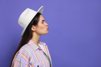 Photo of Beautiful young woman with stylish hat giving kiss on purple background. Space for text